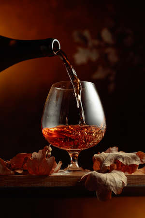 Brandy is poured from a bottle into a glass. A snifter of brandy on an old oak table with dried-up...