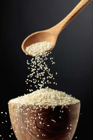 Grains of sesame are poured with a wooden spoon in the bowl. White sesame on a dark background. Copy space.の写真素材