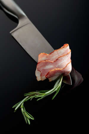 Bacon with rosemary on a kitchen knife. Black reflective background.の写真素材
