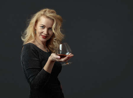 An attractive mature woman in a black evening dress is tasting cognac. Beautiful forty years blonde...
