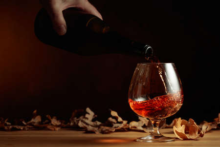 Brandy is poured from a bottle into a glass. A snifter of brandy on an old oak table with dried-up oak leaves. Copy space.の写真素材