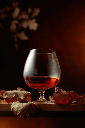 Snifter of brandy on an old oak table with dried-up oak leaves. Copy space.