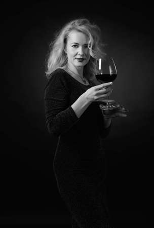 Attractive middle age woman in black evening dress with glass of red wine. Happy forty years blonde on a black background. Black and white.の写真素材