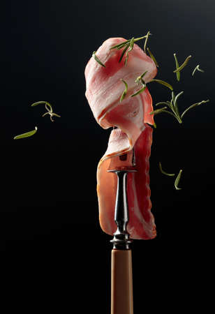 Bacon and rosemary. Smoked bacon on a fork and falling rosemary leaves.の写真素材