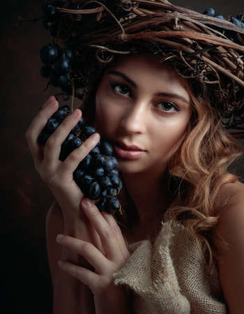 A beautiful blonde girl with long wavy hair holds a grape cluster in her hand. A wreath of vines with blue grapes on the head.の写真素材