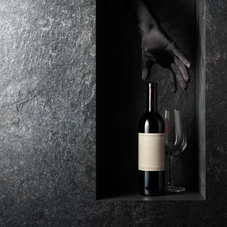 A woman's hand in a black glove reaches for a bottle of red wine with an old empty label. A concept...