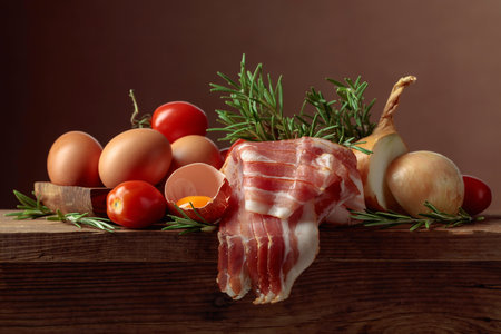 Smoked bacon with eggs, tomatoes, onions, and rosemary on an old wooden table.の写真素材