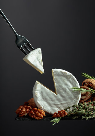 Brie cheese with walnuts, thyme, and rosemary on a black background. Brie cheese and fork with...