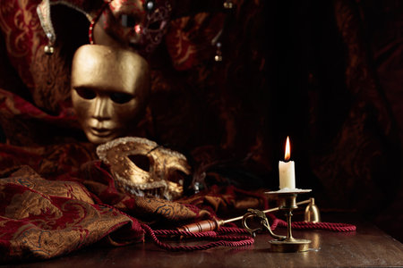 Burning candle in an old brass candlestick and carnival masks on an old wooden table.の写真素材
