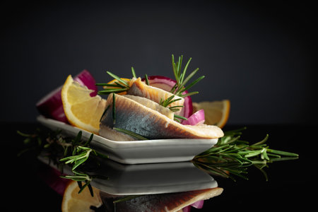 Herring fillet in oil with lemon, red onion, and rosemary. White dish with herring on a black reflective background.の写真素材