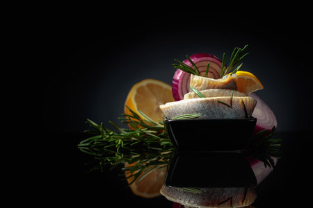 Herring fillet in oil with lemon, red onion, and rosemary. Black dish with herring on a black reflective background. Copy space.の写真素材