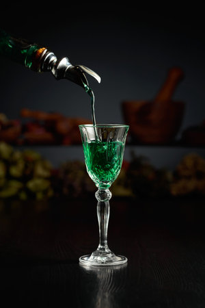 Green herbal liquor is poured from a vintage bottle into a glass. In the background dried herbs and old kitchen utensils.の写真素材