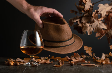 Snifter of brandy on a stone table with dried-up oak leaves and a man's hat.