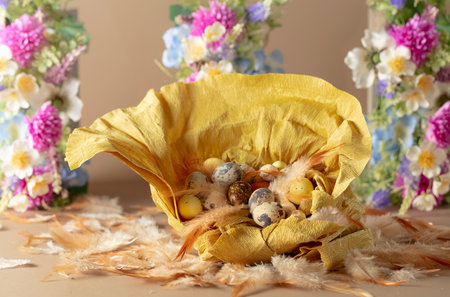 Crepe paper nest with easter eggs, feathers, and spring flowers on a beige background. Focus on a...