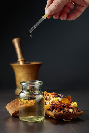 Dropping essential oil or herbal tincture into a glass bottle. Mix of dried healthy medicinal herbs and healing plants on a black background. Copy space.の写真素材