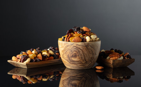 Mix of various nuts and raisins in wooden dish on a black reflective background.