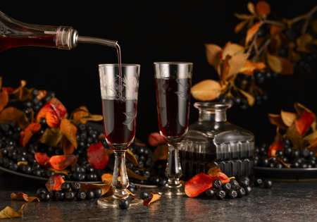 Black chokeberry liqueur and fresh berries on a black stone table. Sweet flavor drink is poured from a bottle into a glass.の写真素材