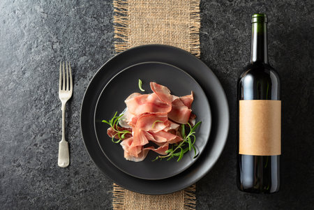 Bottle of red wine and prosciutto on a black plate. On a bottle empty label. Top view.の写真素材
