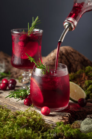 Cranberry cocktail with ice, lime, and rosemary on an old wooden board. Liquor is poured into a...