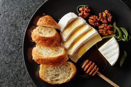 Camembert cream cheese with honey, croissants, and walnuts on a black plate. Top view.