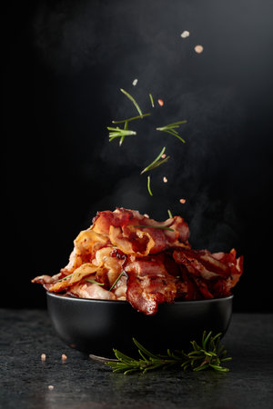 Fried bacon in a black dish, sprinkled with rosemary and pink salt, against a black background with copy space.の写真素材