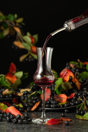 Black chokeberry liqueur and fresh berries on a black stone table. Sweet flavor drink is poured from a bottle into a glass.の写真素材