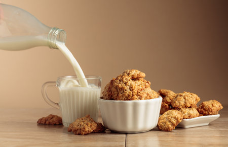 Oatmeal cookies and milk sit on a kitchen table. Milk is poured from a bottle into a glass mug. Beige background with copy space.の写真素材