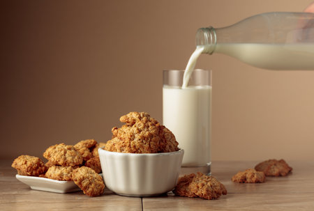 Oatmeal cookies and milk sit on a kitchen table. Milk is poured from a bottle into a glass. Beige background with copy space.の写真素材