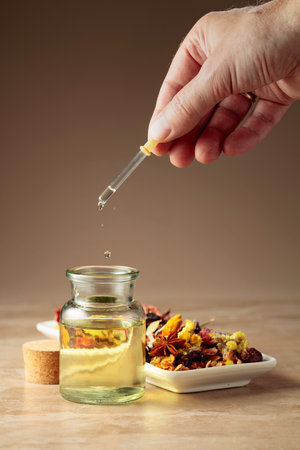 Dropping essential oil or herbal tincture into a glass bottle. Mix of dried healthy medicinal herbs and healing plants on a beige background. Copy space.の写真素材