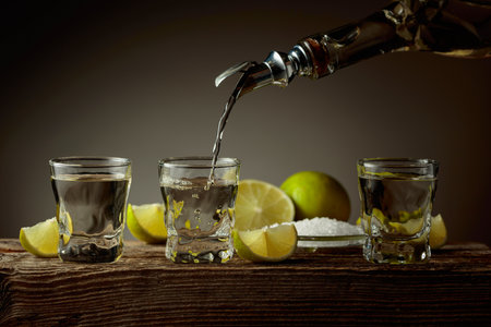 Gold tequila with salt and lime slices on an old wooden board. Tequila is poured from a bottle into a glass.の写真素材