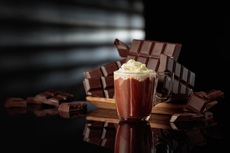 A mug of hot chocolate with airy whipped cream against broken bitter chocolate bars. The black glossy background adds depth and reflection. Copy space.の写真素材