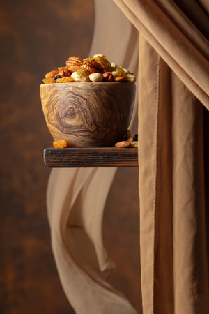 The mix of various nuts and raisins in a wooden bowl on a brown background. In the background there is a beige curtain fluttering in the wind. Copy space.の写真素材