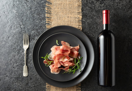 Bottle of red wine and prosciutto on a black plate. Top view. Copy space.の写真素材