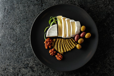 Creamy Camembert with savory olives, fragrant rosemary, and crunchy walnuts on a black plate. A harmonious blend of flavors in an elegant presentation. Top view.の写真素材