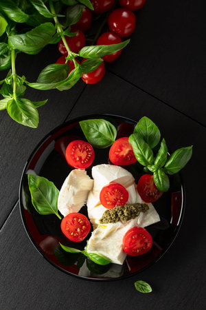 Mozzarella with fresh basil, juicy cherry tomatoes, and vibrant pesto sauce on a black plate. A classic taste of Italy, beautifully arranged. Top view.の写真素材
