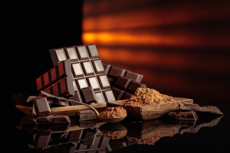 Broken bitter chocolate bars and wooden dish with cocoa powder. The black glossy background adds depth. Focus on a spoon. Copy space.の写真素材