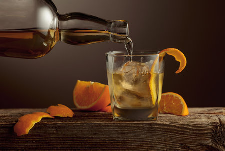 Old Fashioned cocktail with ice and orange peel on an old wooden table. Whiskey is poured from a bottle into a glass with natural ice.の写真素材