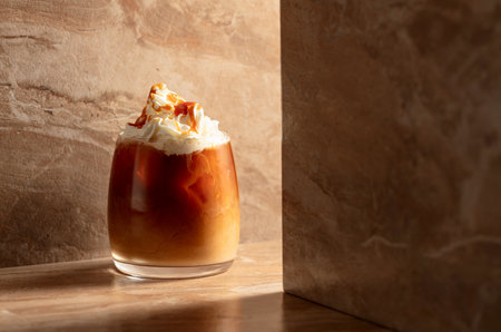 Iced caramel latte topped with whipped cream and caramel sauce, refreshing and sweet coffee drink on a beige ceramic table.の写真素材