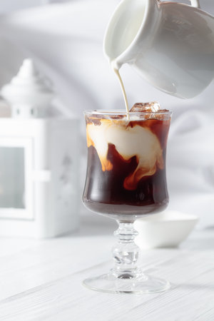 Cream is poured into a glass with iced coffee. Chilled coffee with natural ice on a white wooden table.の写真素材
