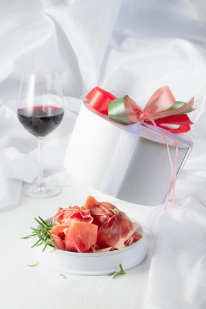 Prosciutto with rosemary in gift box and glass of red wine on a white wooden table. Concept of traditional Italian cuisine.の写真素材