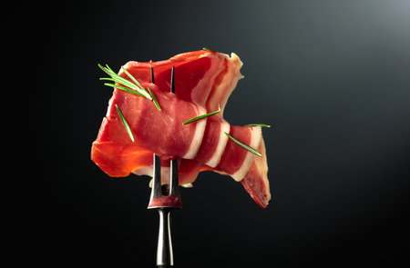 Sliced prosciutto with rosemary on a fork, black background, copy space.の写真素材
