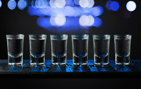 Alcoholic shots of tequila or strong drink in small wet glasses on a black background. Blue backlight. Copy space.の写真素材