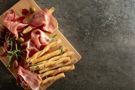 Prosciutto with bread sticks, olives, and rosemary on a wooden cutting board. Traditional Italian snacks on a stone table.の写真素材
