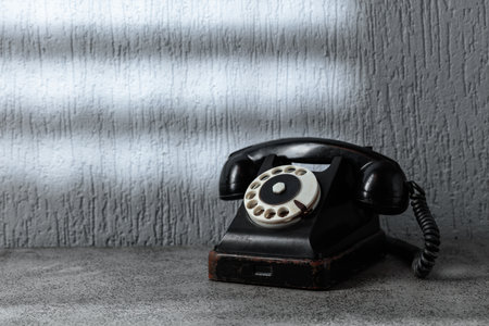 Vintage black telephone with rotary dial on gray stone table. A beam of light falls on the gray wall through a window with a grille. The concept of hope and expectation. Copy space.の写真素材