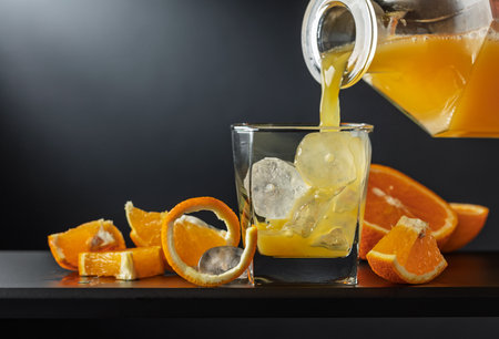 Glass of orange juice with ice on a black background. Juice is poured from a decanter onto a glass with ice. Refreshing drink with fruit slices.の写真素材