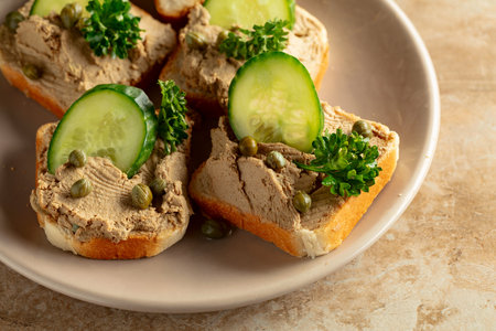 Toasts with pate on a beige plate. Open sandwiches with pate, fresh cucumber, capers, and parsley.の写真素材