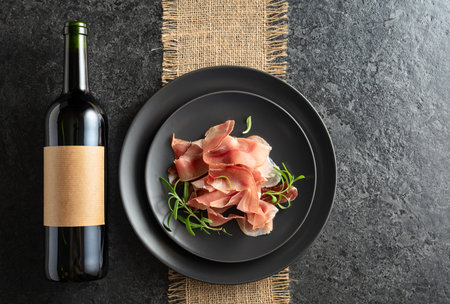 Bottle of red wine and prosciutto on a black plate. On a bottle empty label. Top view.の写真素材