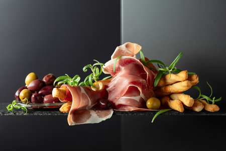 Prosciutto with bread sticks, olives, and rosemary on a black background. Traditional Italian snacks.の写真素材