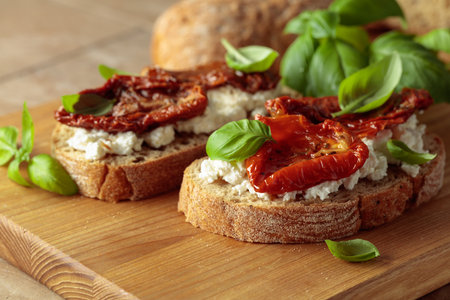 Ciabatta with sun dried tomatoes in olive oil, ricotta, and basil. Traditional Italian snack on a wooden cutting board.の写真素材