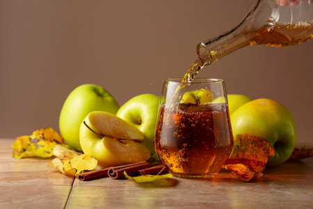Apple juice is poured from a bottle into a glass. On a wooden table ripe apples, cinnamon sticks, and dried-up leaves. Beige background with copy space.の写真素材